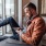 a man sits on a chair and rests his foot on a balcony railing as he looks at his phone in his room